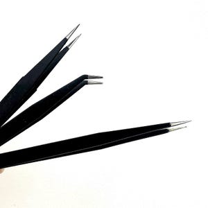 Puede incluir: Tres pinzas negras con puntas plateadas se muestran sobre un fondo blanco. Las pinzas tienen diferentes estilos de punta, incluyendo puntas puntiagudas y anguladas. Las herramientas parecen nuevas y sin usar.
