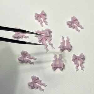 May include: A collection of small, light pink bow-shaped embellishments. Some bows have long, flowing ribbons. A pair of black tweezers holds one bow. The bows are scattered on a white surface.