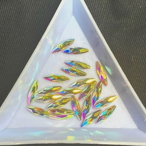 Può includere: Una collezione di strass iridescenti e allungati in un vassoio triangolare di plastica bianca. Gli strass hanno un design sfaccettato, che riflette uno spettro di colori tra cui rosa, giallo e blu. Il vassoio è su una superficie scura.