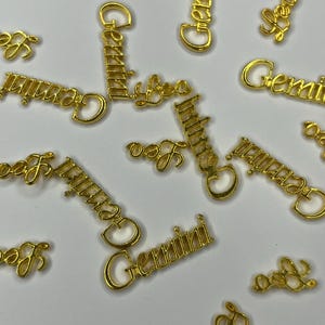 Puede incluir: Amuletos de metal dorado esparcidos sobre una superficie blanca. Los amuletos presentan las palabras "Gemini" y "Leo" en una fuente cursiva. Los amuletos son probablemente para la fabricación de joyas o manualidades.