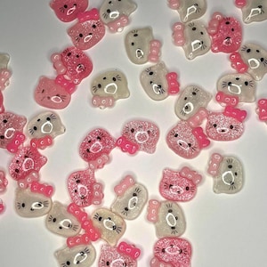 Pode incluir: Uma coleção de pingentes de resina rosa e transparente em forma de gato com laço. Alguns pingentes têm detalhes de glitter. Os pingentes estão espalhados em uma superfície branca.