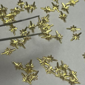 May include: A scattering of small, gold-coloured star-shaped embellishments on a white surface. The stars have multiple points and are likely for crafts or decoration. A pair of tweezers is also visible.
