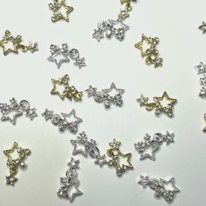 Könnte beinhalten: Nahaufnahme verschiedener sternförmiger Anhänger in Gold- und Silbertönen. Einige Anhänger haben einen einzelnen Stern, während andere mehrere Sterne haben, die mit kleinen Ketten verbunden sind. Die Sterne sind mit kleinen, klaren Kristallen verziert.