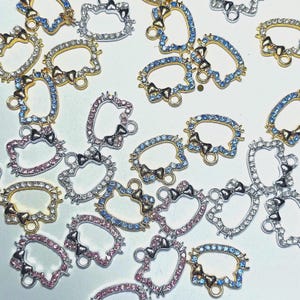 May include: An assortment of rhinestone-studded charms in various colors, including gold, silver, and pink. The charms feature a bow and a cat-like face design, with a small loop at the bottom for attachment. The rhinestones are clear and blue.