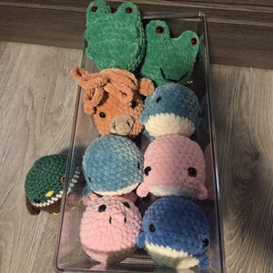 May include: A clear plastic container filled with various crocheted plush toys. The toys include green frogs, an orange cow, blue and pink whales, a blue hippo, and a green turtle. The toys are soft and have black safety eyes.