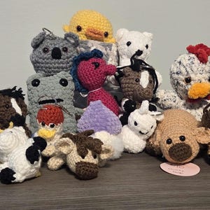 May include: A collection of handmade crochet animal toys, including a koala, duck, bear, chicken, frog, horse, cow, and whale. The toys are in various colors and sizes, with detailed features.