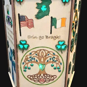 Puede incluir: Una linterna de madera con temática irlandesa con tréboles iluminados, las banderas estadounidense e irlandesa y la frase "Erin go Bragh!". La linterna presenta un diseño de árbol de la vida y el texto "Irish and Proud of our roots."