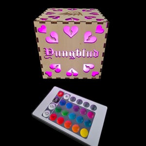 Könnte beinhalten: Eine helle Holzwürfellampe mit lasergeschnittenen, gebrochenen Herzformen und dem Schriftzug "YUNGBLUD", der von innen in hellem Pink leuchtet. Die Lampe wird mit einer rechteckigen Fernbedienung mit verschiedenen Farbtasten geliefert.