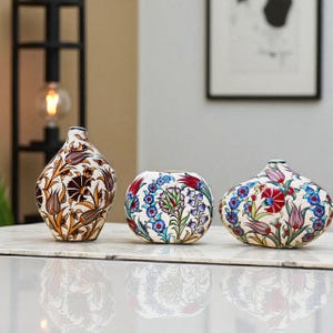 May include: Three ceramic vases with floral patterns. The vases are in different shapes: a tall, teardrop-shaped vase, a round vase, and a flattened, oval vase. The floral designs include red, blue, and brown flowers with green leaves on a cream background.
