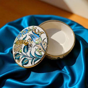 May include: A small, round trinket box with a lid, open to reveal the interior. The lid features a floral design in blue, green, and gold on a white background. The box has a gold rim and sits on a blue satin surface.