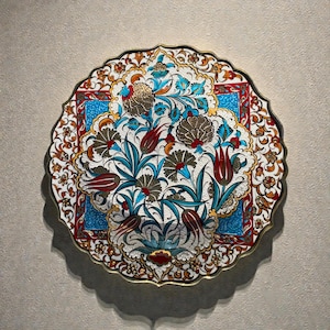 May include: A round, decorative plate with a scalloped edge. The plate features a floral design with red tulips, teal leaves, and gold accents on a white background. The center has a blue square with more floral details.