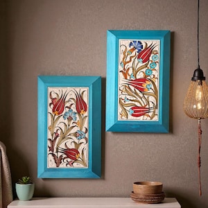 Può includere: Due opere d'arte incorniciate con un design floreale. Ogni pezzo presenta una piastrella rettangolare con una cornice turchese. Le piastrelle mostrano tulipani rossi, fiori blu e foglie verdi su uno sfondo color crema. L'opera d'arte è esposta su un muro marrone.