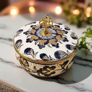 May include: A round trinket box with a lid, featuring a white base and a floral pattern in blue and gold. The lid is adorned with a gold handle. The box has a gold rim and a textured surface.