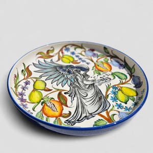 May include: A round ceramic plate with a blue rim, featuring a hand-painted design. The artwork depicts a winged figure amidst colorful fruits and floral elements in shades of yellow, orange, blue, and green. The plate's interior is white, providing a contrasting backdrop.