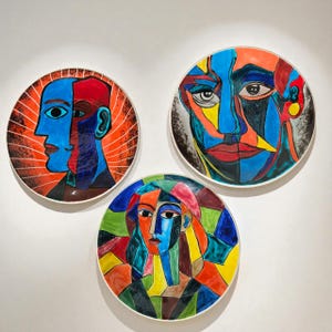 May include: Three decorative ceramic plates with abstract face designs. Each plate features a different colorful geometric portrait. The plates are round with a white border and are mounted on a white wall.