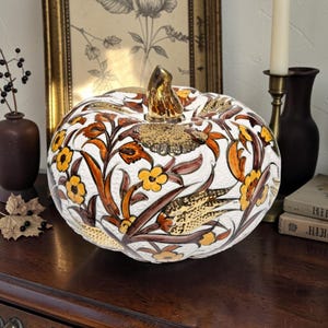 Pode incluir: Uma abóbora decorativa de cerâmica com uma superfície branca texturizada, adornada com padrões florais pintados à mão em laranja e amarelo, caules castanhos e detalhes metálicos dourados. A abóbora é rematada por um caule dourado brilhante.