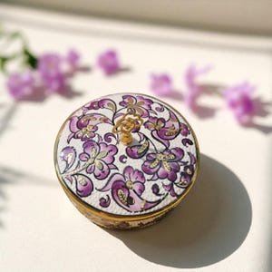 May include: A round trinket box with a white base and a floral pattern in purple and gold. The lid has a gold handle and small, sparkling details. The box is edged with a gold rim. The box is approximately 7.6 cm in diameter.