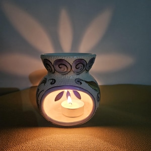 Può includere: Un diffusore di oli essenziali in ceramica bianca con bordo turchese e design a spirale viola. All'interno del diffusore c'è una candela accesa, che proietta un bagliore caldo e ombre a forma di foglia.