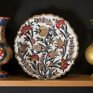 Puede incluir: Un plato decorativo blanco con borde festoneado y borde dorado. El plato presenta un diseño floral con elementos rojos, dorados y negros. Las flores y hojas están dispuestas en un patrón simétrico. El plato se exhibe en una estantería de madera.