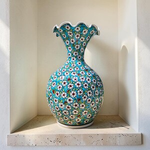 May include: A decorative ceramic vase with a turquoise background and a pattern of small white flowers with red centers. The vase has a ruffled rim and a bulbous body, and is displayed on a stone shelf.