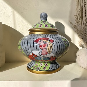 May include: A decorative ceramic jar with a lid. The jar features a hand-painted design of a woman in a red hat, with a striped background. The lid and base are accented with gold trim, and the lid has a green floral pattern.