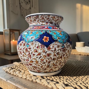 Peut inclure: Un vase décoratif en céramique avec un corps large et un col étroit. Le vase présente un motif floral détaillé en blanc, bleu, orange et rouge. Le vase mesure environ 25 cm de haut et repose sur une base blanche.