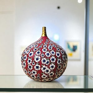 Op de afbeelding: Een decoratieve vaas met een rode achtergrond en een patroon van witte en blauwe bloemen. De vaas heeft een goudkleurige hals en staat op een glazen plank. De vaas is een uniek kunstwerk.