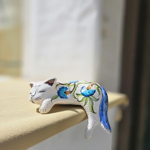May include: A white ceramic cat figurine with blue floral designs and green leaves. The cat is lying down with its tail hanging over the edge of a surface. The cat's eyes are closed, and it has a relaxed expression.