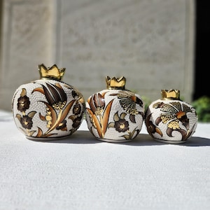 May include: Three decorative ceramic pomegranate-shaped vases. The vases are white with brown and gold floral designs and a textured surface. Each vase has a gold crown-shaped lid. The vases are arranged in a row, decreasing in size.