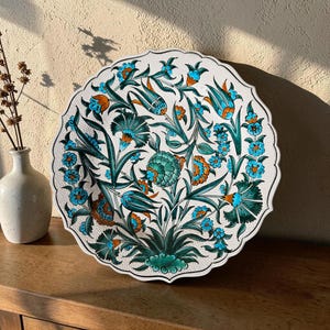 Op de afbeelding: Een decoratieve witte keramische plaat met een geschulpte rand, versierd met een bloemmotief in turquoise, oranje en groen. Het gedetailleerde ontwerp toont bloemen en bladeren, wat een levendige en ingewikkelde esthetiek creëert.