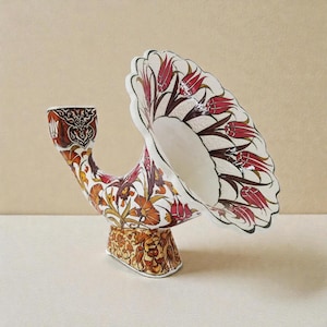 Può includere: Un vaso decorativo in ceramica dalla forma curva unica. Il vaso presenta un interno bianco ed è decorato con un motivo floreale nei toni del rosso, arancione e marrone. La base è rettangolare con un motivo dettagliato.