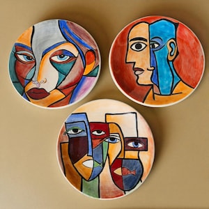 Könnte beinhalten: Drei dekorative Keramikplatten mit abstrakten Gesichtsdesigns. Jede Platte zeigt eine einzigartige, farbenfrohe geometrische Komposition mit kräftigen Linien und einer Vielzahl von Farben, darunter Rot, Blau, Orange und Grau. Die Teller sind rund und scheinen handbemalt zu sein.