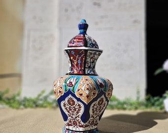 May include: A decorative ceramic vase with a lid. The vase features a vibrant design with blue, red, yellow, and white floral patterns. The base is square, and the lid has a small knob on top.