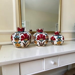 Peut inclure: Trois vases en céramique en forme de grenade de tailles variées, présentant une surface blanche texturée ornée de motifs floraux peints à la main en rouge, jaune et bleu sarcelle, surmontés d'un bord rouge foncé et d'accents dorés.