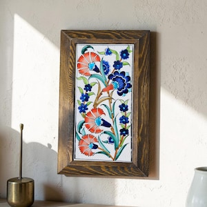 May include: A framed ceramic tile with a floral pattern. The artwork displays bright orange and blue flowers with green leaves on a white backdrop. The frame is made of dark wood, giving the piece a rustic appearance.