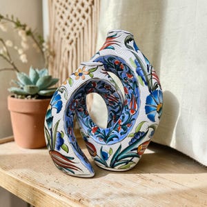 Op de afbeelding: Een witte keramische vaas met een uniek, open ontwerp. De vaas is versierd met een kleurrijk bloemmotief in blauw, rood en oranje. De vaas is ongeveer 25 cm hoog en staat op een houten oppervlak.