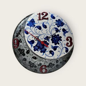 May include: Round wall clock with a white face featuring blue floral designs. The clock has a black and gray patterned border. The numbers 12, 3, 6, and 9 are in a dark red color. The clock hands are black and red.