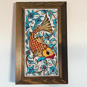 May include: A framed ceramic tile featuring a stylized orange and red fish swimming amidst blue floral patterns. The artwork is set against a white background and framed in a dark brown wooden frame, creating a decorative wall art piece.