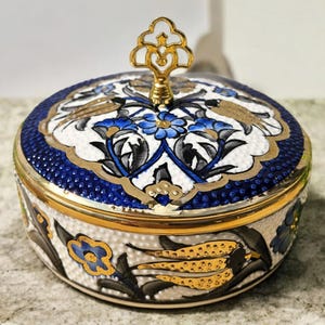 May include: A round ceramic trinket box with a lid. The box has a white base with blue and gold floral patterns. The lid features a blue dotted border and a gold handle. The design is ornate and decorative.