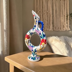 May include: A white ceramic ring-shaped vase featuring a central circular opening, a tall slender neck, and a small handle. The surface is decorated with colorful floral patterns in shades of red, blue, purple, green, and yellow throughout.