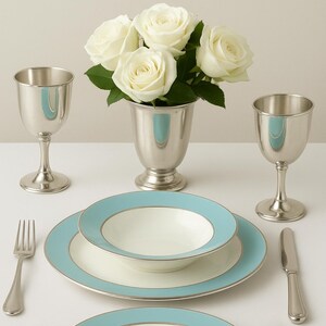 May include: Elegant table setting featuring a bouquet of white roses in a silver vase, flanked by two silver goblets. The setting includes white plates and bowls with a light blue border and silver trim, along with silverware.