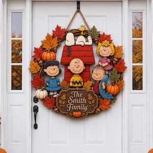 May include: A circular autumn wreath featuring Peanuts characters including Snoopy on a red doghouse, Charlie Brown, Lucy, Linus, and Sally. The wreath is decorated with orange and white pumpkins, acorns, and red, orange, and green maple leaves. A central brown plaque displays the text The Smith Family.