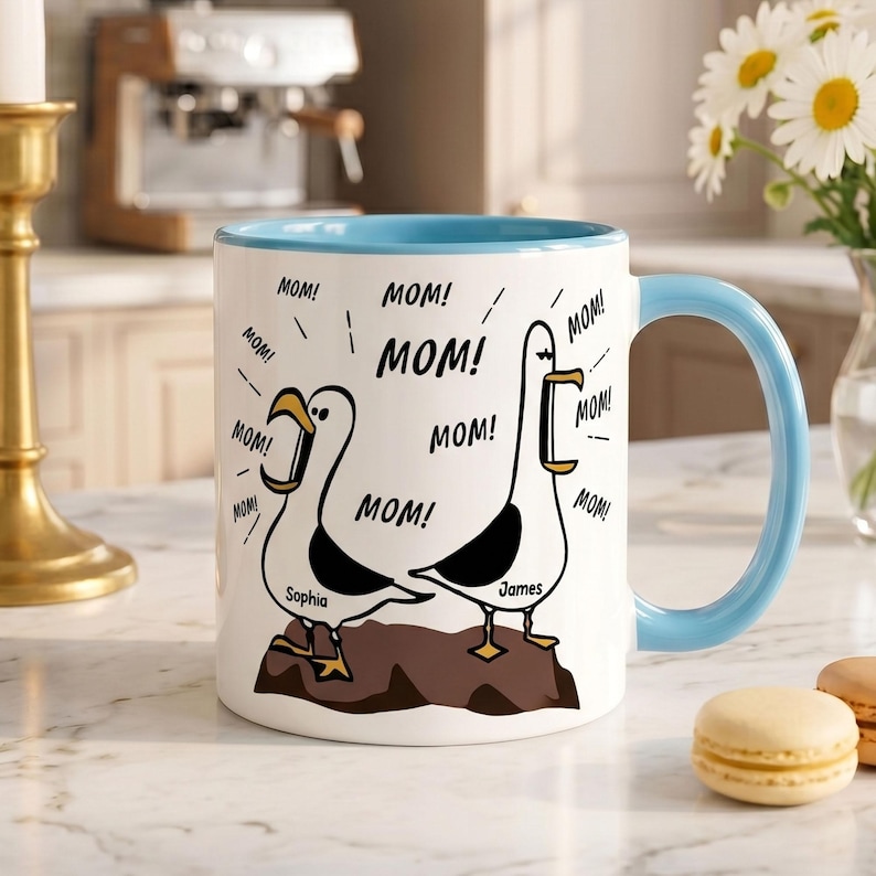 Peut inclure: Mug en c&eacute;ramique blanche avec une anse et un bord int&eacute;rieur bleu clair. Le mug pr&eacute;sente des mouettes de dessin anim&eacute; avec le mot "MOM!" r&eacute;p&eacute;t&eacute; autour d'elles. Les noms "Sophia" et "James" sont imprim&eacute;s sur le mug.