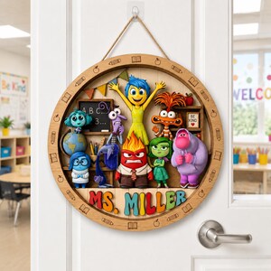 May include: A circular wooden wall sign featuring colorful 3D figures of characters from the movie Inside Out, including Joy, Sadness, Anger, Disgust, Fear, and Bing Bong. The sign includes the text MS. MILLER and hangs from a jute rope.