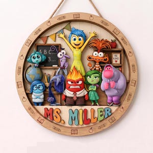 May include: A circular wooden wall sign featuring colorful 3D figures of characters from Inside Out in a classroom setting. The frame is engraved with pencils, books, and apples. The text MS. MILLER is displayed in multi-colored 3D letters.