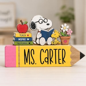 May include: A wooden pencil-shaped desk sign with a yellow body and pink eraser, featuring the text MS. CARTER. It includes cutouts of Snoopy reading a blue book, Woodstock, a stack of books labeled TEACH, INSPIRE, ENCOURAGE, and a potted flower.