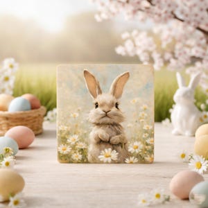 May include: A square decorative tile featuring a watercolor painting of a bunny in a field of daisies. The bunny is light brown with long ears and is standing upright. Pastel colored Easter eggs and a white ceramic bunny are in the background.