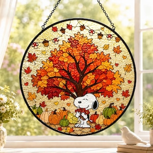 May include: A circular stained glass suncatcher featuring Snoopy holding a bouquet of red flowers under a large autumn tree with orange and yellow leaves. The scene includes pumpkins, gourds, and a string of mushrooms and acorns against a textured background.