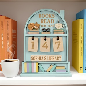 May include: A light blue wooden bookshelf-shaped display with the text "BOOKS READ THIS YEAR" and "SOPHIA'S LIBRARY". It features a sleeping fox, glasses, and a coffee cup. The display shows the number 147.