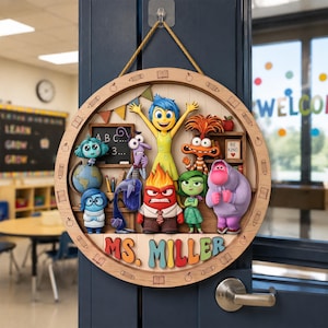 May include: A circular wooden classroom door sign featuring colorful 3D-style characters from Inside Out, including Joy, Sadness, Anger, Disgust, Fear, Bing Bong, and Anxiety. The sign displays the text MS. MILLER and is suspended by a gold rope.